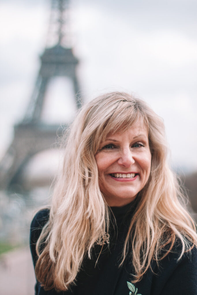 The picture shows Christina Mantel, MA smiling in front of the Eiffel Tower in Paris.